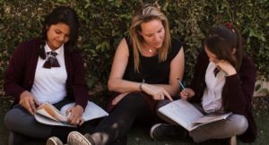 Aspire principal sitting in between two students, sitting on grass and helping both with their assignments. Students are holding paper and pencils in their laps.